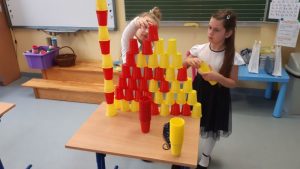Dwie uczennice układają wieżę z żółtych i czerwonych plastikowych kubeczków.